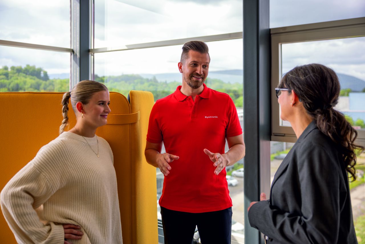 man in red Bystronic polo short talks to two women